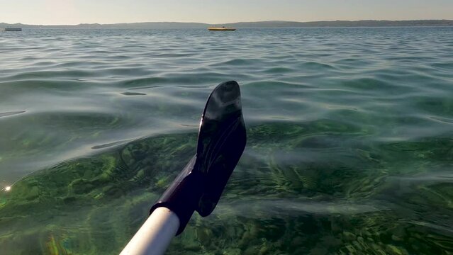 Kayak Paddle Skimming the Transparent Lake Waters. The water is so clear the bottom of the lake can be seen. A buoy and various boats can be seen in the distance. 