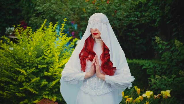 Fantasy woman in white vintage dress holy virgin prays face is hidden by silk veil. Red-haired girl nun queen. Medieval princess bride in garden green trees flowers. Mystical image sexy lady vampire