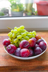 green grapes and plums in a plate on a wooden board, windowsill, concept of fresh fruits and healthy food