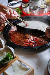 A view hand dipping raw meat slices into the broth bowl, seen with of all the elements of a hot pot restaurant meal.