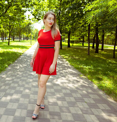 A young woman in a red dress stands on the road in the summer park near the trees. Woman portrait
