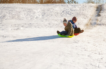 Fototapeta na wymiar In winter, a boy and a girl ride down a hill on a plastic sled., brother and sister ride plastic plates from the mountain.