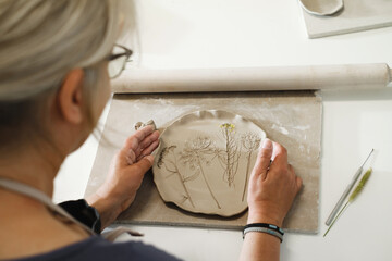 Woman rolling clay, making ceramic plate in studio with floral pattern. Handmade creative work. Pottery workshop for adults. Top view on plate