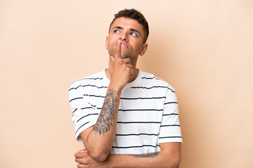 Young Brazilian man isolated on beige background having doubts while looking up