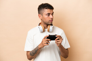 Young Brazilian man playing with a video game controller isolated on beige background looking to the side