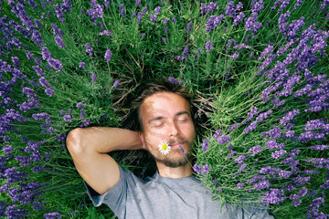 Portrait of young handsome bearded man lying among lavender flowers in blossom field. Happy smiling guy relax on the grass on sunny summer day. Self care mind body and soul.