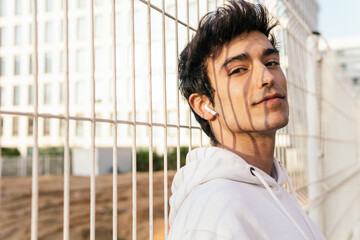 Stylish man standing near metal grid with wireless earphones in city
