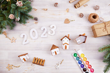 Happy New Year 2023 and Merry Christmas greeting card. Homemade toy wooden houses, white numbers 2023, fir branches, gift wrapping paper, paints on a light background.