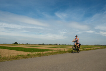 Obraz premium A young lady riding her bicycle on a cycle path through very typical Dutch countryside in the Netherlands on a clear sunny summer day. Example of uniform precision farming can be seen behind