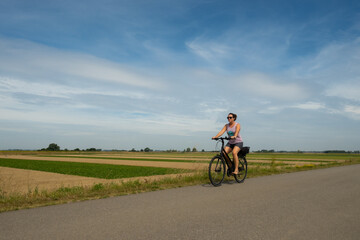 Fototapeta na wymiar A young lady riding her bicycle on a cycle path through very typical Dutch countryside in the Netherlands on a clear sunny summer day. Example of uniform precision farming can be seen behind