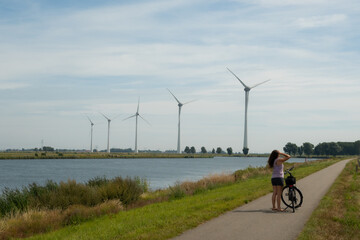 A female cyclist has taken a break from her journey in front of a group of 5 very big wind power generating turbines tower over farmland in the Dutch countryside in the Netherlands. 