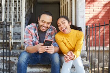 Excited ethnic couple surfing internet on smartphone on stairs outdoors