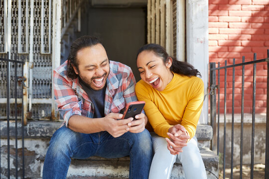 Excited ethnic couple surfing internet on smartphone on stairs outdoors