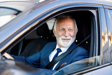 An old executive drives himself to work. A smiling happy senior businessman sits in his car with hands on the steering wheel and looks through the window.