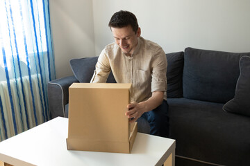 Happy young man unpacking delivery box, sitting on couch by coffee table, home interior. Satisfied customer cheerful man opening package gadgets on Internet, copy space.