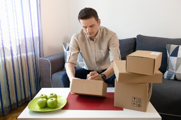 Young male online seller and start up small business owner writing address on cardboard box and packing parcels for delivery at home.