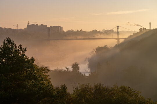 Scenic sunrise in Zhytomyr city. Morning fog over valley of Teteriv river. High pedestrian cable bridge and residential buildings on background, Ukraine