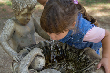 Image of an adorable little girl drinking water from a decorated fountain in a public park. Games...