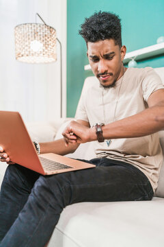 Confused black man looking at wristwatch while using laptop