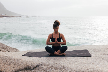 Fototapeta na wymiar Fit woman with crossed legs meditating while sitting on mat by ocean