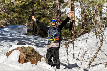 Happy snowboarder man with arms raised on background of forest in the mountains. Sunny warm...