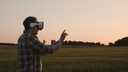 Farmer in virtual reality helmet in front of a sunset agricultural landscape. Man in a countryside field. Country life, food production, farming and technology concept