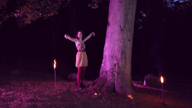 Beautiful young and smiling woman with pumpkin halloween makeup emerging from behind a tree, with a small pumpkin candle in hand, welcomes you, waving hello come here to the halloween event