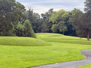 Walking path and curved earthworks prehistoric Great Serpent Mound. Body of snake effigy visible along landscape with woods and dramatic sky.