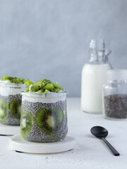 Chia pudding with coconut milk, greek jogurt and shape heart kiwi on a marble coasters. Glass bottle with milk, jar with chia seeds and black matte teaspoon on white table. Grey background. Copy space