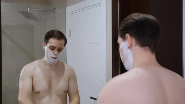 Serious young guy shaving his beard in a bathroom by razer.