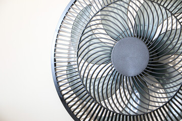 Close up of spinning black electric fan showing its rotating vanes and blades, on right of frame, against white wall background, necessary appliance for summer heat waves and rising temperatures