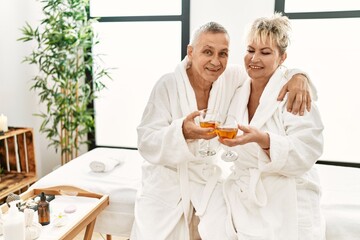 Senior caucasian couple smiling happy toasting with champagne sitting on massage table at beauty center.