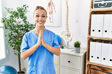 Young caucasian woman working at pain recovery clinic praying with hands together asking for forgiveness smiling confident.