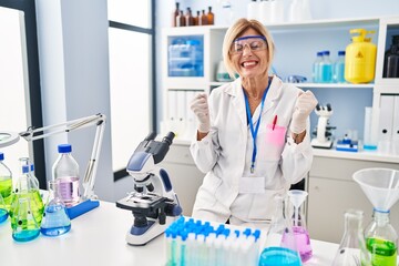 Middle age blonde woman working at scientist laboratory excited for success with arms raised and eyes closed celebrating victory smiling. winner concept.