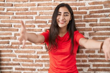 Young teenager girl standing over bricks wall looking at the camera smiling with open arms for hug. cheerful expression embracing happiness.