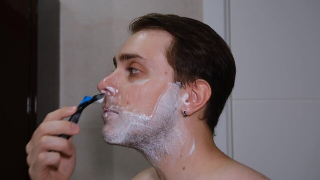 Serious young guy shaving his beard in a bathroom by razer.