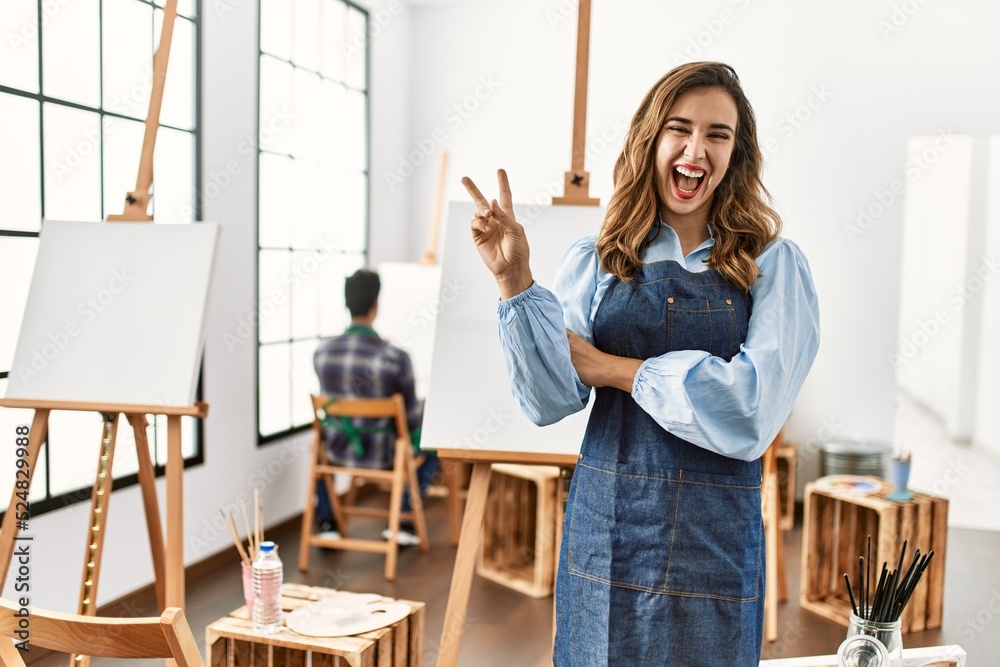 Poster young artist woman at art studio smiling with happy face winking at the camera doing victory sign. n