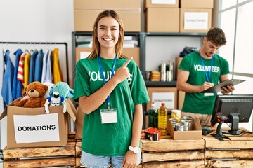 Young blonde girl wearing volunteer t shirt at donation stand cheerful with a smile on face pointing with hand and finger up to the side with happy and natural expression