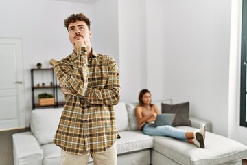 Fototapeta premium Young handsome man at the living room with girlfriend serious face thinking about question with hand on chin, thoughtful about confusing idea
