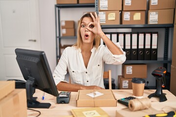Young blonde woman working at small business ecommerce doing ok gesture shocked with surprised face, eye looking through fingers. unbelieving expression.