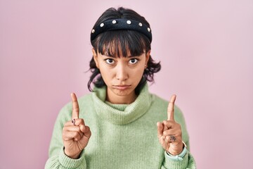 Young beautiful woman standing over pink background pointing up looking sad and upset, indicating direction with fingers, unhappy and depressed.