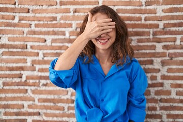 Beautiful brunette woman standing over bricks wall smiling and laughing with hand on face covering eyes for surprise. blind concept.