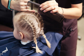 Barber woman make fashionable hairstyle for cute little blond girl child in modern barbershop. Hair salon, Hairdresser makes hairdo braids pigtail for young baby in barber shop. Copy space - obrazy, fototapety, plakaty