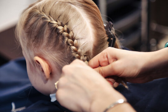 Hair salon, hairdresser makes hairdo braids for young baby in barber shop. Barber woman make fashionable hairstyle for cute little blond girl child in modern barbershop. Concept hairstyle and beauty