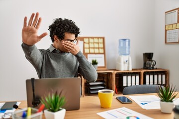 Young hispanic man wearing business style sitting on desk at office covering eyes with hands and doing stop gesture with sad and fear expression. embarrassed and negative concept.