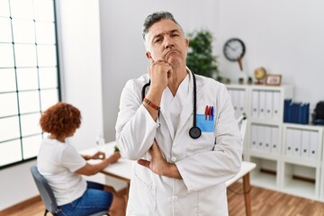 Middle age doctor man at the clinic with a patient with hand on chin thinking about question,...