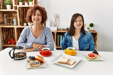 Family of mother and down syndrome daughter sitting at home eating breakfast winking looking at the camera with sexy expression, cheerful and happy face.