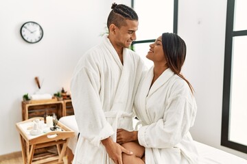 Young latin couple wearing bathrobe hugging and sitting on massage table at beauty center.