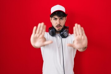 Hispanic man with beard wearing gamer hat and headphones doing stop gesture with hands palms, angry and frustration expression