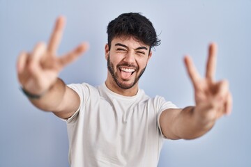 Hispanic man with beard standing over white background smiling with tongue out showing fingers of both hands doing victory sign. number two.